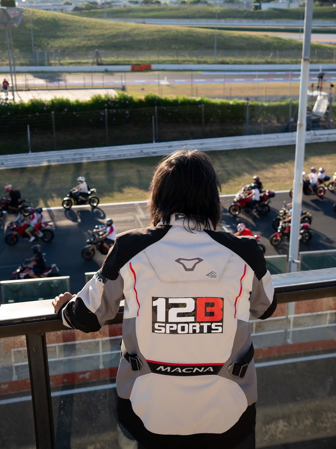 Person wearing a 12B SPORTS motorcycle jacket overlooking riders on a racetrack during an international motorsport experience.