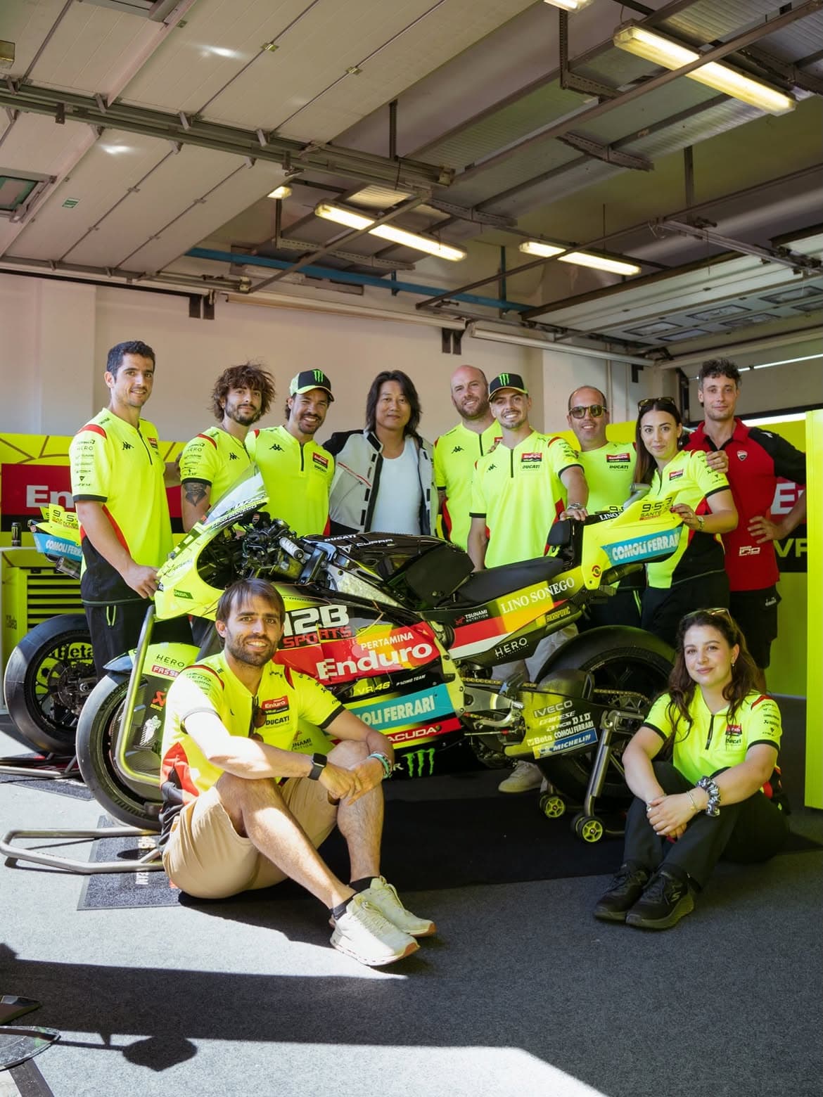 Motorsport team and guests posing around a racing motorcycle featuring prominent 12B SPORTS branding inside the team garage.